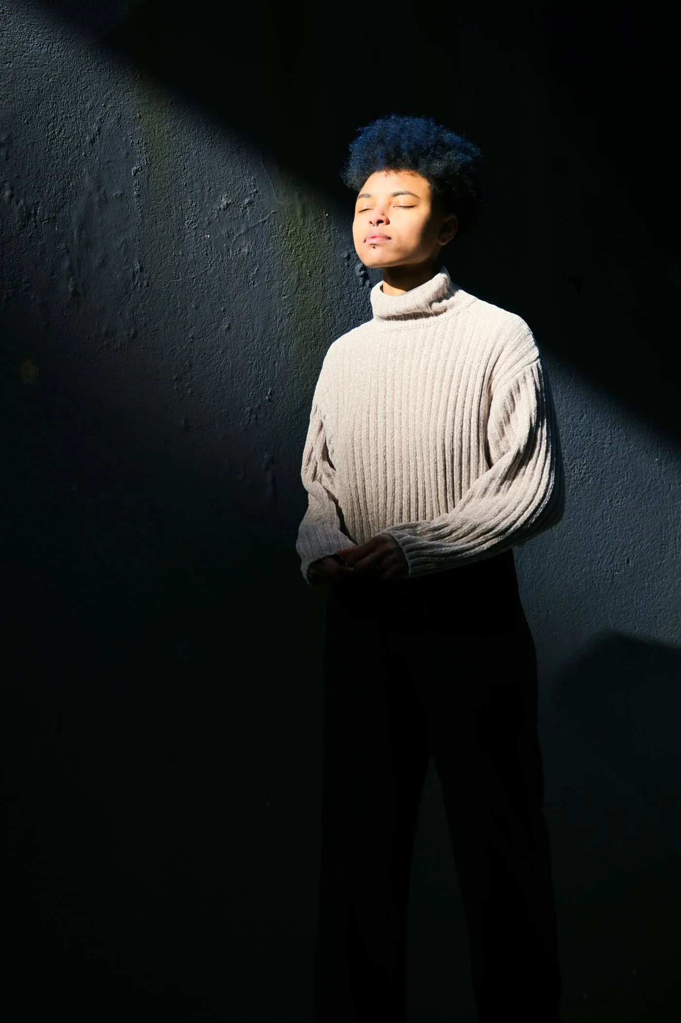 A person in a grey high-neck knit standing in a patch of sunlight against a dark wall.