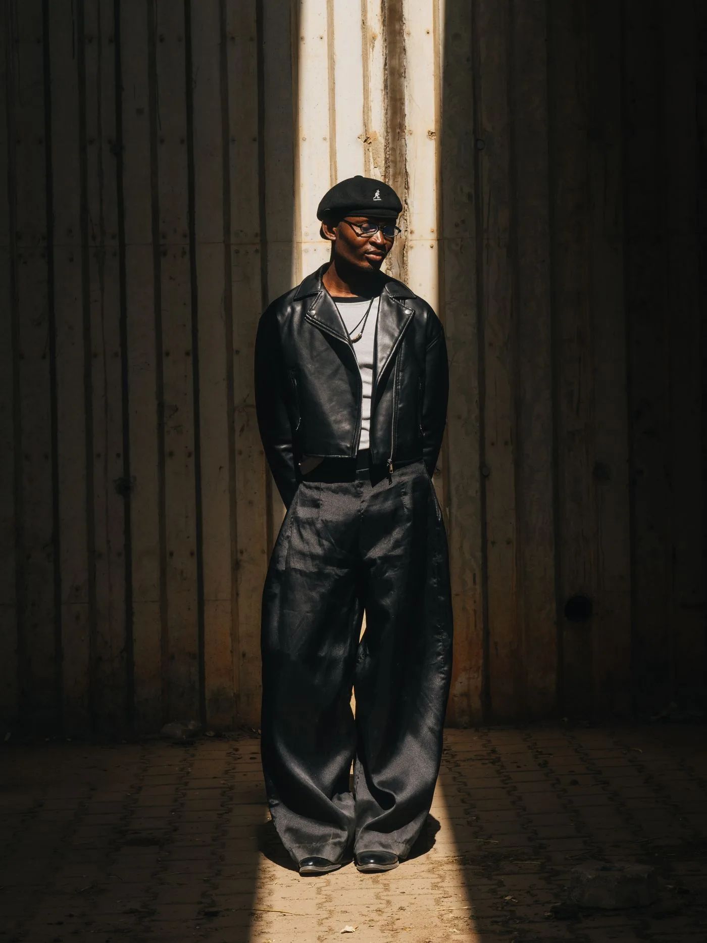 A person in a dark jacket and wide trousers standing in sunlight.