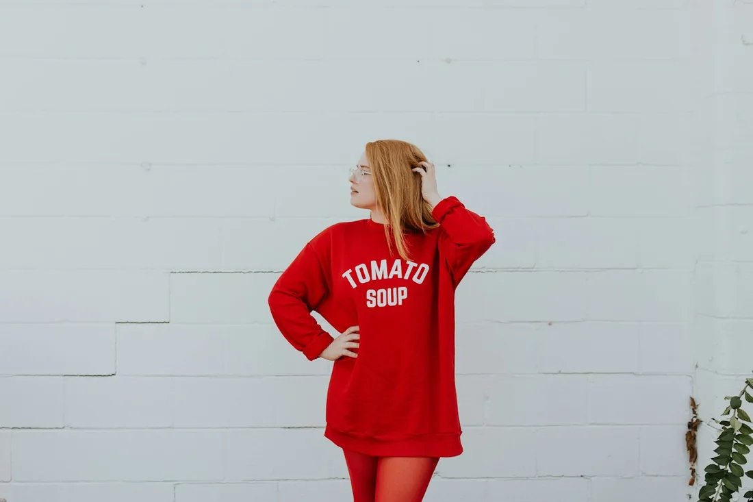 A woman wearing a red graphic sweatshirt beside a pale wall.